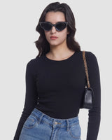 A woman wearing a fitted black long-sleeve top and blue jeans, styled with black cat-eye sunglasses and a small black shoulder bag with a gold chain strap, against a light gray studio background.