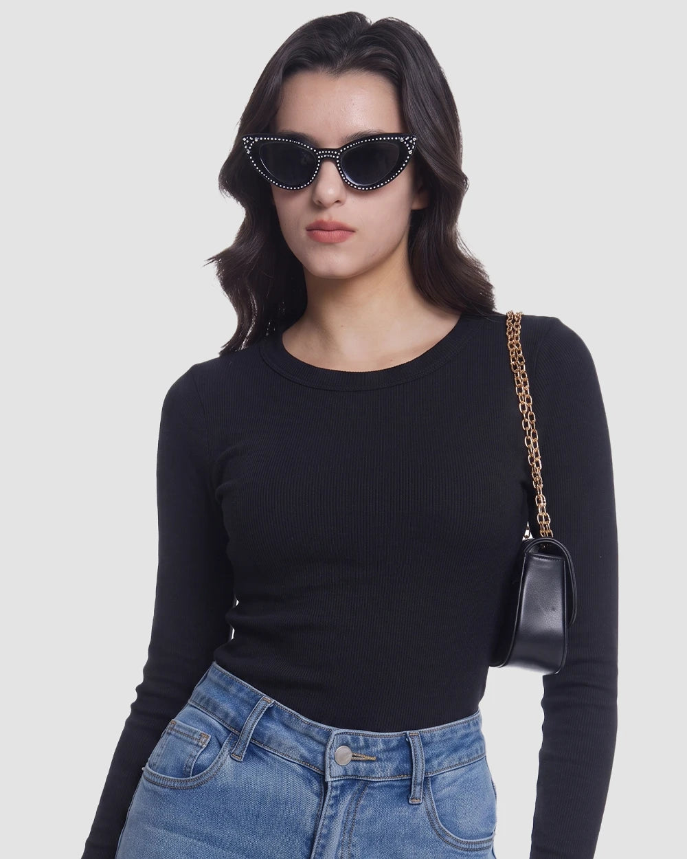 A woman wearing a fitted black long-sleeve top and blue jeans, styled with black cat-eye sunglasses and a small black shoulder bag with a gold chain strap, against a light gray studio background.
