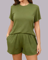 A woman wearing a matching olive green lounge set consisting of a short-sleeve crewneck t-shirt and high-waisted shorts, shown against a solid gray studio background.