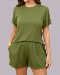 A woman wearing a matching olive green lounge set consisting of a short-sleeve crewneck t-shirt and high-waisted shorts, shown against a solid gray studio background.