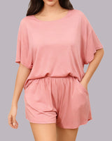 A woman wearing a dusty pink two-piece lounge set consisting of a short-sleeve top and matching shorts, shown against a solid gray background.
