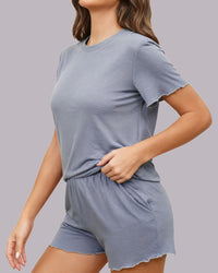 A woman wearing a light blue-gray short-sleeve t-shirt and matching shorts with scalloped edges, shown from the neck down against a solid gray studio background.