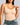 A woman wearing a light beige seamless shapewear bodysuit with underwire cups and thin shoulder straps, paired with blue jeans, against a solid gray background.