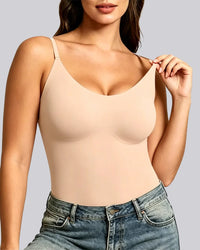 A woman wearing a light beige seamless shapewear bodysuit with underwire cups and thin shoulder straps, paired with blue jeans, against a solid gray background.