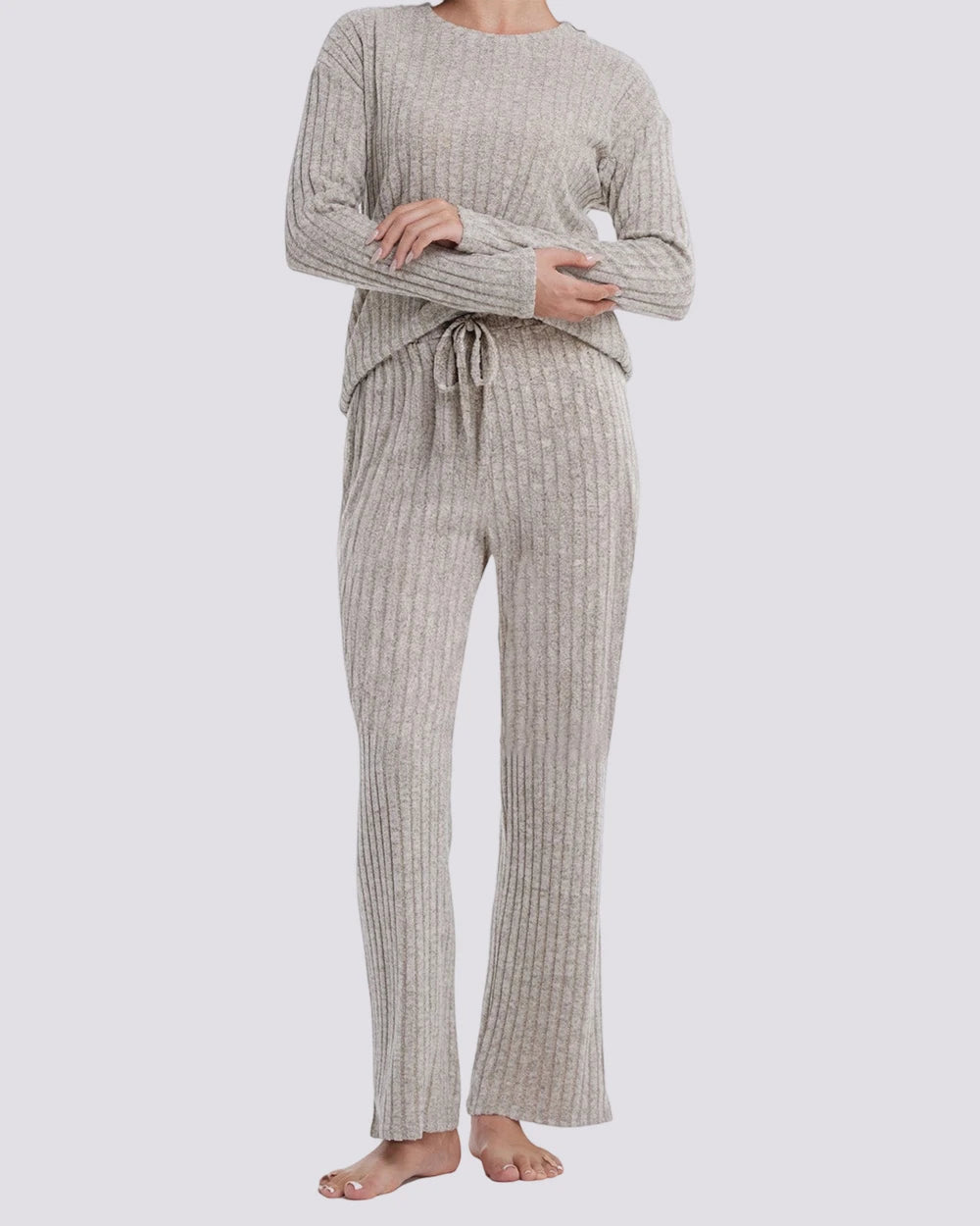 A woman modeling a ribbed knit two-piece loungewear set in a light oatmeal beige color, featuring a long-sleeve top and matching drawstring pants, against a solid light gray background.