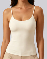 A woman wearing a fitted spaghetti-strap camisole tank top in warm off-white/cream, paired with light beige pants, against a plain light gray studio background.