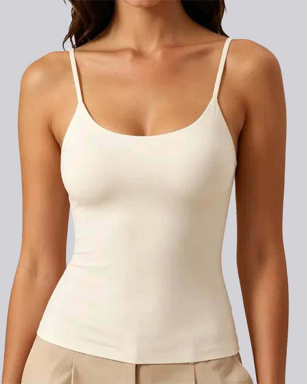 A woman wearing a fitted spaghetti-strap camisole tank top in warm off-white/cream, paired with light beige pants, against a plain light gray studio background.
