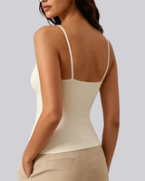 A woman shown from the back wearing a fitted spaghetti-strap camisole top in a warm off-white/cream color, paired with beige pants, against a solid light gray background.