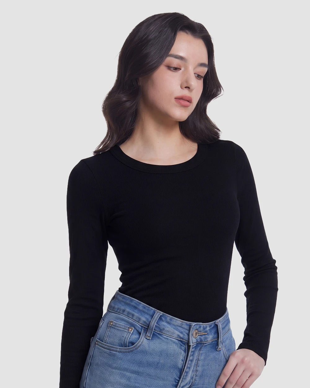 A woman wearing a fitted black long-sleeve crewneck top and blue jeans, photographed against a plain light gray studio background.