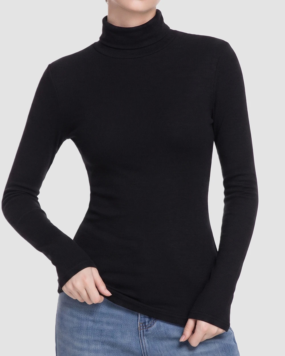 A woman wearing a fitted black long-sleeve turtleneck top with blue jeans, shown from the neck down against a light gray studio background.