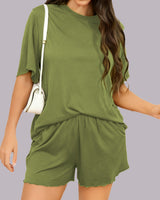 A woman wearing a matching olive green short-sleeve t-shirt and shorts set, styled with a white shoulder bag, against a solid light gray background.