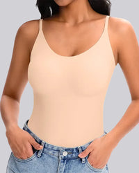 A woman wearing a light beige, skin-toned camisole tank top with thin straps, paired with blue jeans, shown against a solid gray background.