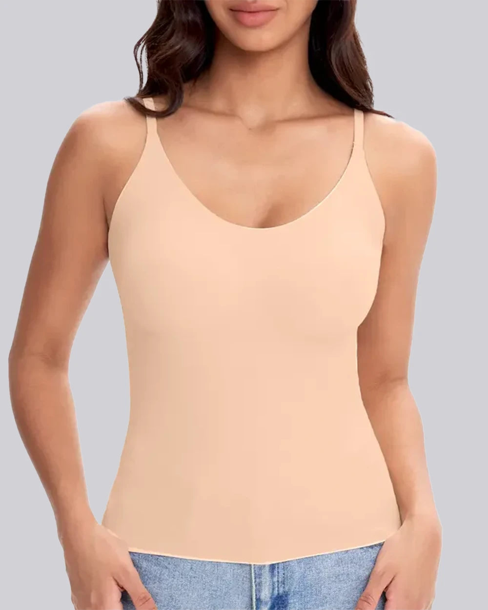 A woman wearing a light peach-beige spaghetti-strap tank top with a scoop neckline, paired with blue jeans, shown against a solid gray background.