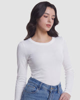 A woman wearing a fitted white long-sleeve crewneck top and blue jeans, photographed against a plain light gray studio background.