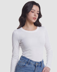 A woman wearing a fitted white long-sleeve crewneck top and blue jeans, photographed against a plain light gray studio background.