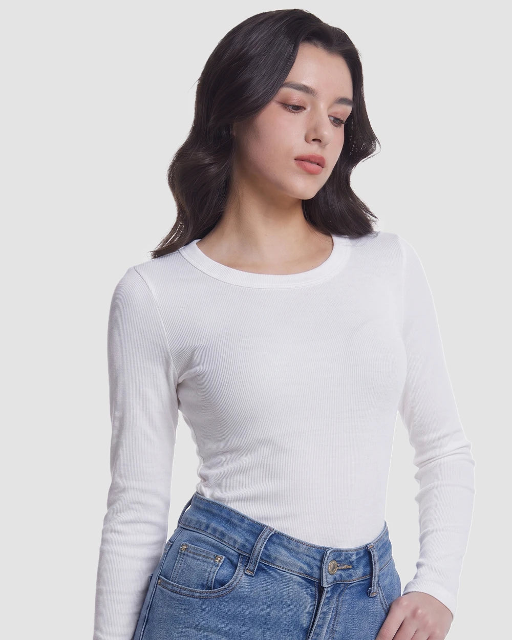 A woman wearing a fitted white long-sleeve crewneck top and blue jeans, photographed against a plain light gray studio background.