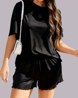 A woman wearing a black short-sleeve t-shirt and matching black shorts with a scalloped hem, carrying a small white shoulder bag against a solid gray background.