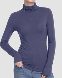 A woman wearing a fitted long-sleeve turtleneck top in a muted blue-purple color, paired with blue jeans, shown against a light gray studio background.