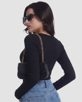 A woman shown from the back wearing a fitted black long-sleeve top, light blue jeans, black sunglasses, and carrying a small black shoulder bag with a gold chain strap against a light gray studio background.