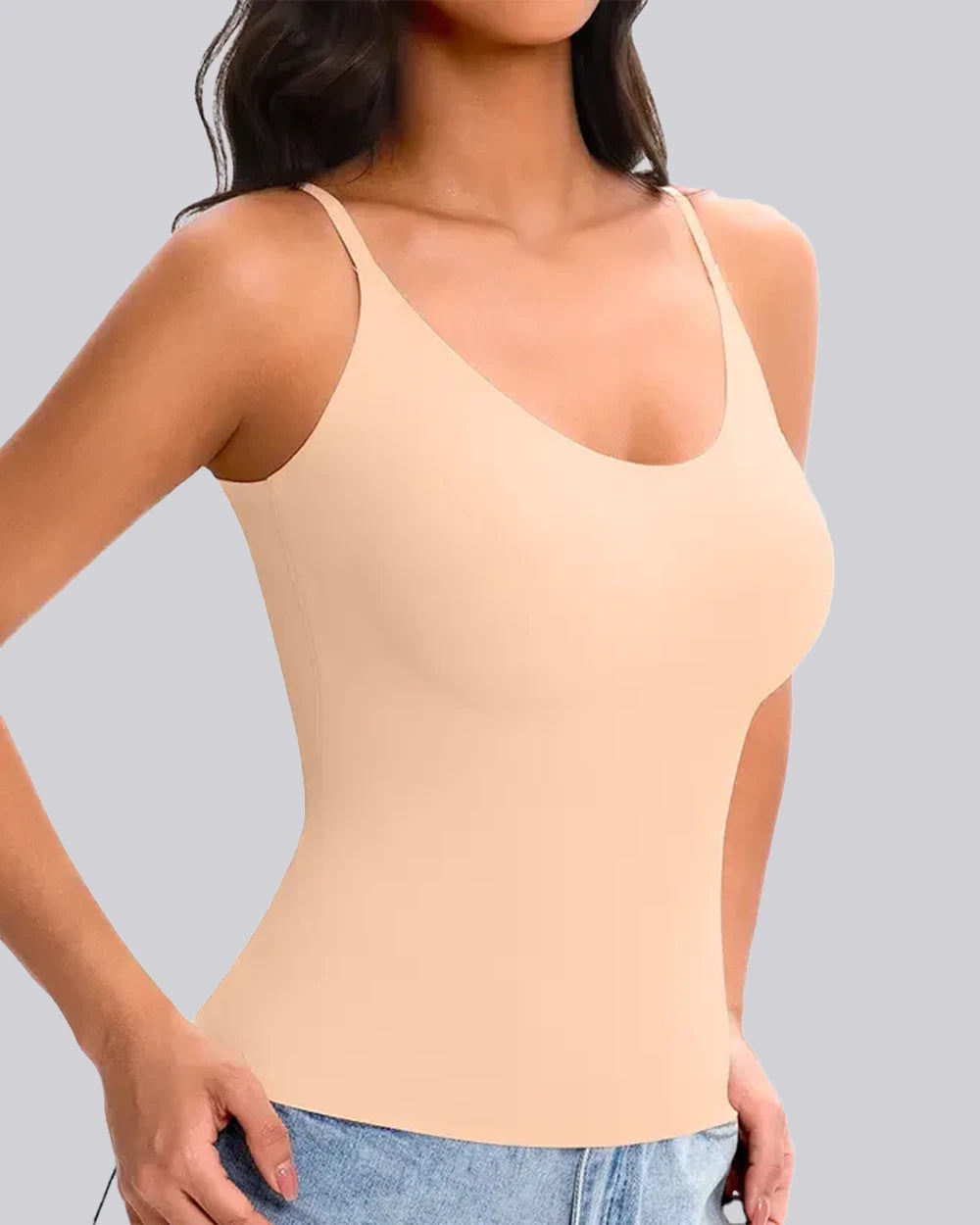 A woman wearing a light peach-beige spaghetti-strap camisole tank top paired with light-wash blue jeans, shown from the neck down against a solid gray background.