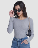 A woman wearing a fitted light gray long-sleeve crewneck top, blue jeans, black cat-eye sunglasses, and a small black shoulder bag with a gold chain strap, posed against a light gray studio background.