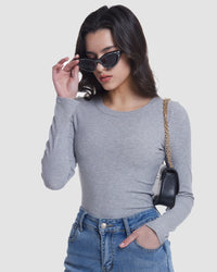 A woman wearing a fitted light gray long-sleeve crewneck top, blue jeans, black cat-eye sunglasses, and a small black shoulder bag with a gold chain strap, posed against a light gray studio background.
