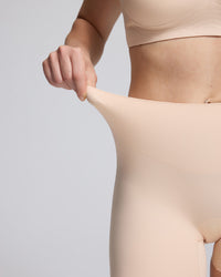 Close-up of a model wearing a light beige high-waisted shapewear brief and pulling the waistband slightly outward with one hand to demonstrate stretch, against a solid light gray background.
