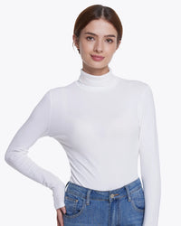 A woman modeling a fitted white long-sleeve mock neck top paired with blue jeans against a plain light gray studio background.