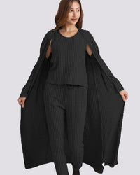 A woman modeling a black ribbed knit three-piece outfit consisting of a sleeveless top with shoulder cutouts, matching wide-leg pants, and a long open-front cardigan, shown against a solid light gray background.