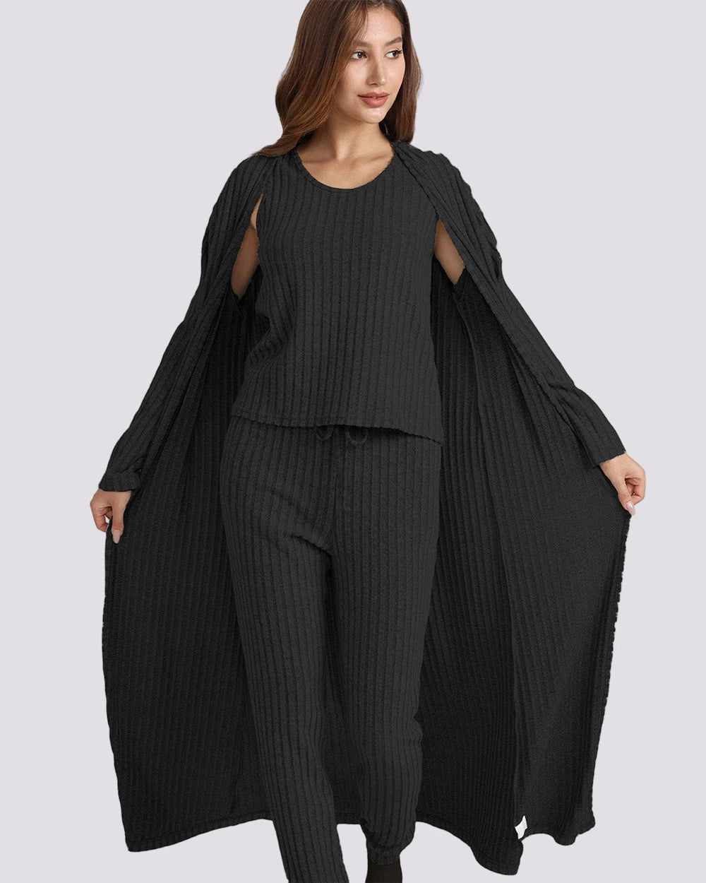 A woman modeling a black ribbed knit three-piece outfit consisting of a sleeveless top with shoulder cutouts, matching wide-leg pants, and a long open-front cardigan, shown against a solid light gray background.