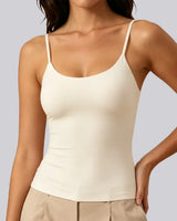 A woman wearing a fitted cream-colored spaghetti-strap camisole tank top paired with beige pants, shown from the neck down against a solid light gray background.