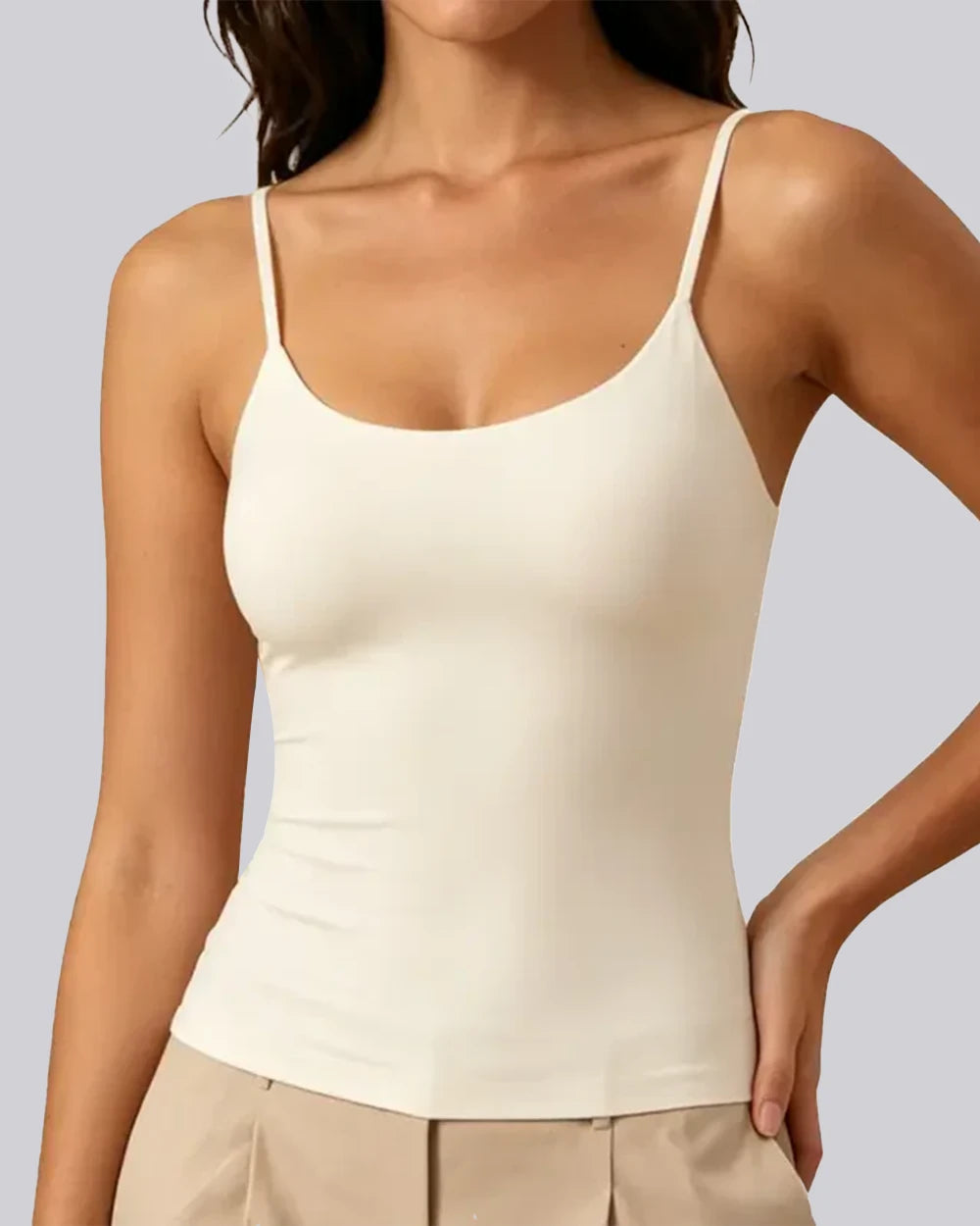 A woman wearing a fitted cream-colored spaghetti-strap camisole tank top paired with beige pants, shown from the neck down against a solid light gray background.