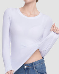A woman wearing a white long-sleeve crewneck top, pulling the fabric slightly to show its stretch and semi-sheer texture, paired with blue jeans against a light gray background.
