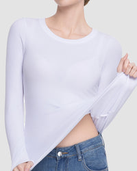 A woman wearing a white long-sleeve crewneck top, pulling the fabric slightly to show its stretch and semi-sheer texture, paired with blue jeans against a light gray background.