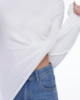 A close-up of a person wearing a white long-sleeve top and blue jeans, pulling the side of the shirt outward to show stretch and fit.