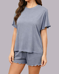 A woman wearing a matching two-piece loungewear set consisting of a short-sleeve t-shirt and shorts, both in a heathered dusty blue-gray color with scalloped edges, against a solid gray studio background.