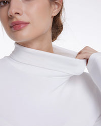 Close-up of a woman wearing a white long-sleeve turtleneck top, pulling the collar slightly outward with one hand against a plain white background.