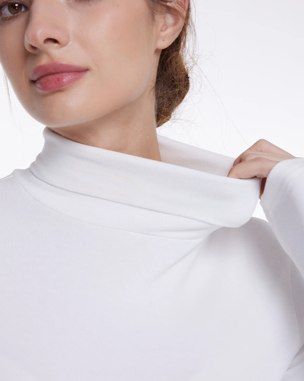 Close-up of a woman wearing a white long-sleeve turtleneck top, pulling the collar slightly outward with one hand against a plain white background.