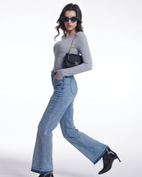 A woman modeling a light gray long-sleeve top, light blue wide-leg jeans, black sunglasses, and black ankle boots, carrying a small black crossbody bag with a gold chain strap against a plain white studio background.