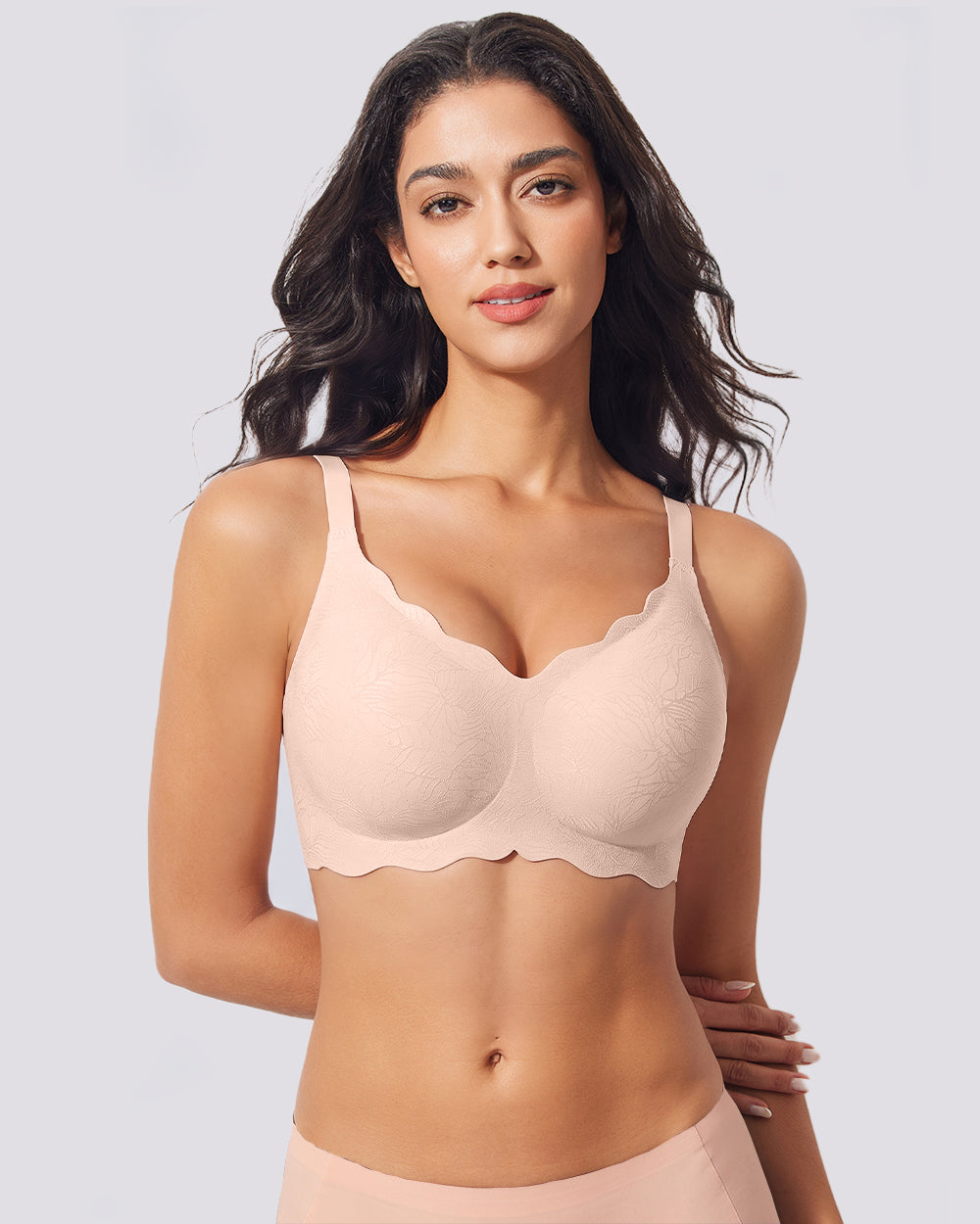 A woman modeling a light peach-beige wireless bra with scalloped edges against a solid light gray background.