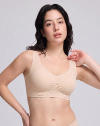 A woman modeling a light beige wireless bra with a gathered sweetheart neckline, photographed against a solid light gray studio background.