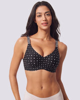 A woman modeling a black underwire bra with VRCOMFY black polka dots and gray patterned shoulder straps, set against a solid light gray background.