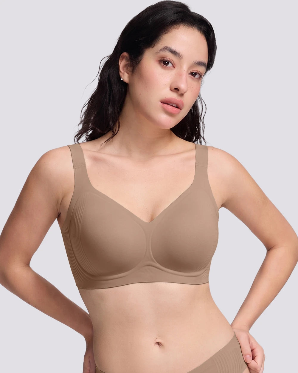 A woman modeling a seamless, VRCOMFY wireless bra in a solid taupe-beige color, shown from the chest down against a light gray studio background.