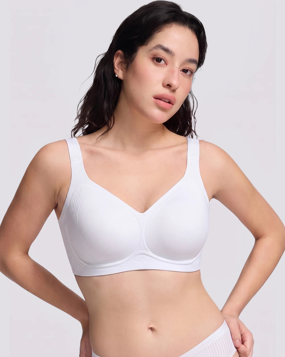A woman modeling a white wireless bra with a V-neckline and wide shoulder straps, photographed against a light gray studio background.