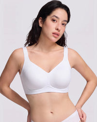 A woman modeling a white wireless bra with a V-neckline and wide shoulder straps, photographed against a light gray studio background.