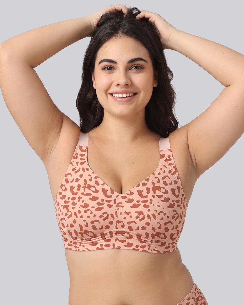 A woman wearing a peach and rust leopard-print bralette, posing with her hands in her hair against a solid gray background.