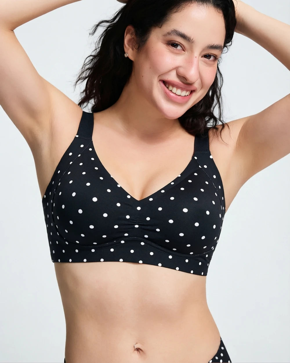 A woman wearing a black V-neck bralette with white polka dots, posing against a light gray studio background.