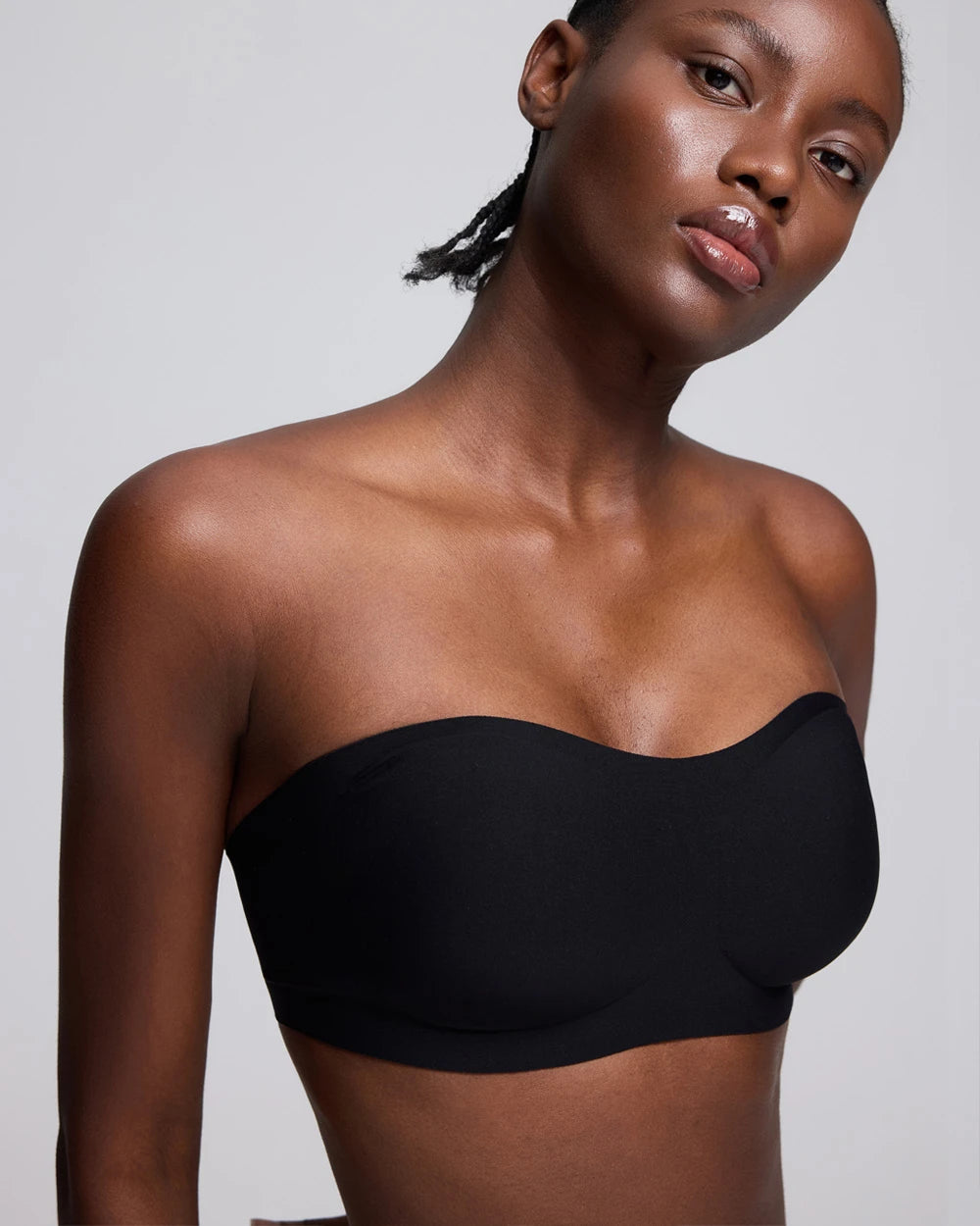 A close-up of a model wearing a black strapless bandeau-style bra or underwire bra against a light gray studio background.