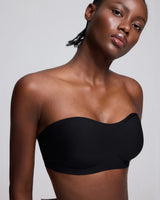 A close-up of a model wearing a black strapless bandeau-style bra or underwire bra against a light gray studio background.