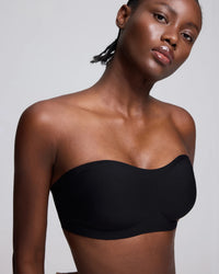 A close-up of a model wearing a black strapless bandeau-style bra or underwire bra against a light gray studio background.